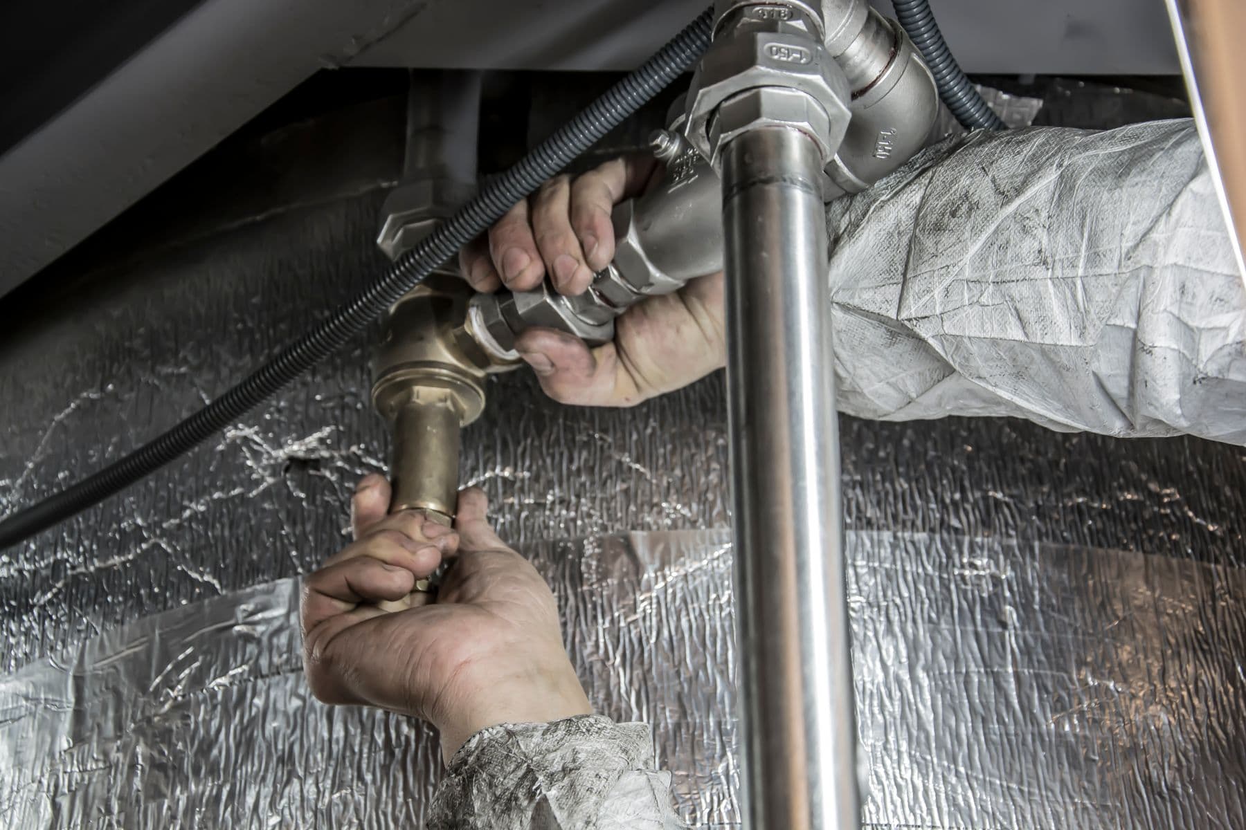 A plumber's gloved hands fitting a stainless steel pipe connection under a fixture